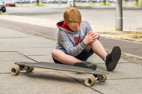 Child holding knee in pain sitting on sidewalk next to a skateboard
