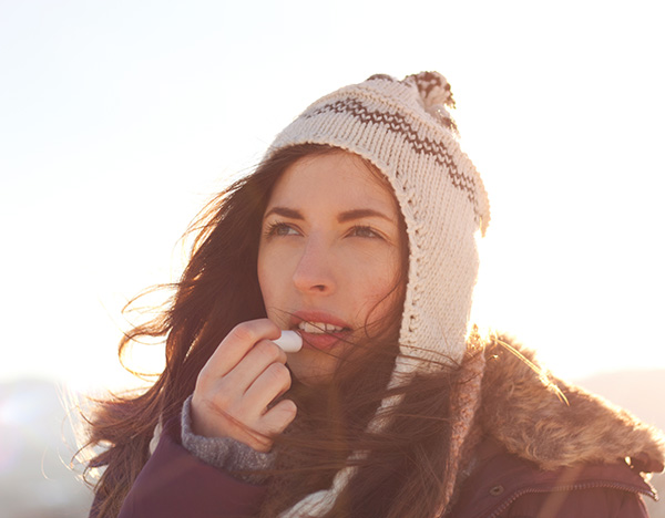 woman in winter coat and hat during golden hour applying chapstick to lips