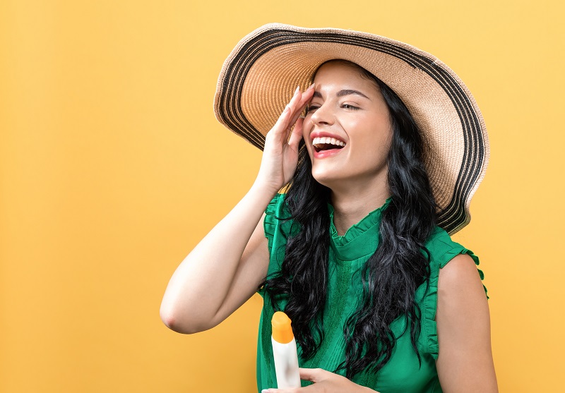 woman in big straw sun hat and green sun dress laughing while applying sunscreen to her face
