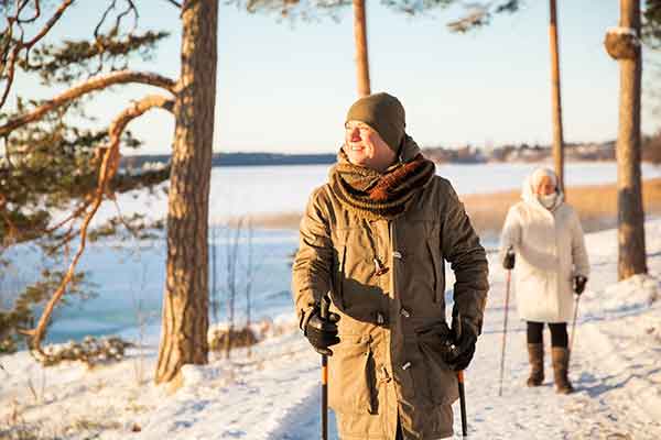 man and woman bundled up in hats and long coats on a snow walk next to a lake and tress