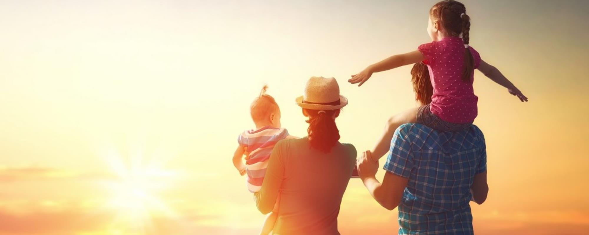 Family of four, with a young child in the mothers arms, and the other child on fathers shoulders, all looking at a vivid yellow and orange sunset