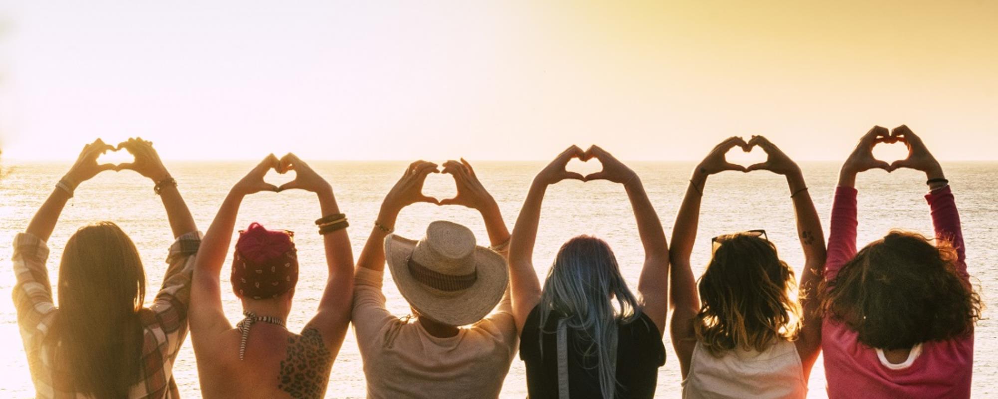 Group of 6 adults sitting side by side, arms raised above head with hands making heart shape against an ocean sunset